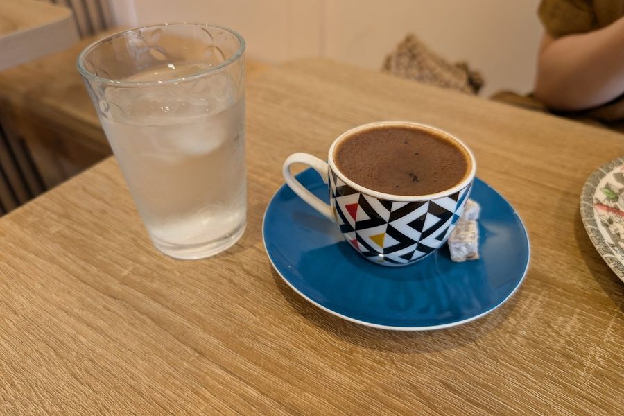 A cup of Turkish coffee with a piece of Turkish delight on a blue saucer, beside a glass of water on a wooden table