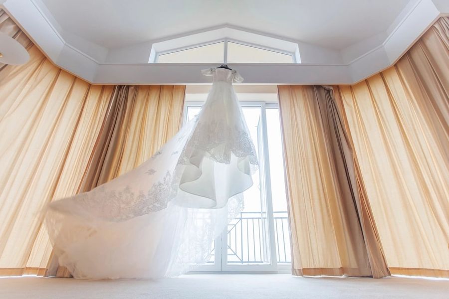 A wedding dress on a hanger in front of a bright window with floor-length curtains