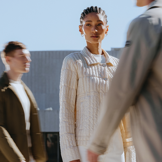 Kvinne med fletter og øreringer ser rett frem mot kamera, mens en uskarp person står i forgrunnen til høyre. Klar blå himmel i bakgrunnen gir bildet et rent og minimalistisk uttrykk.