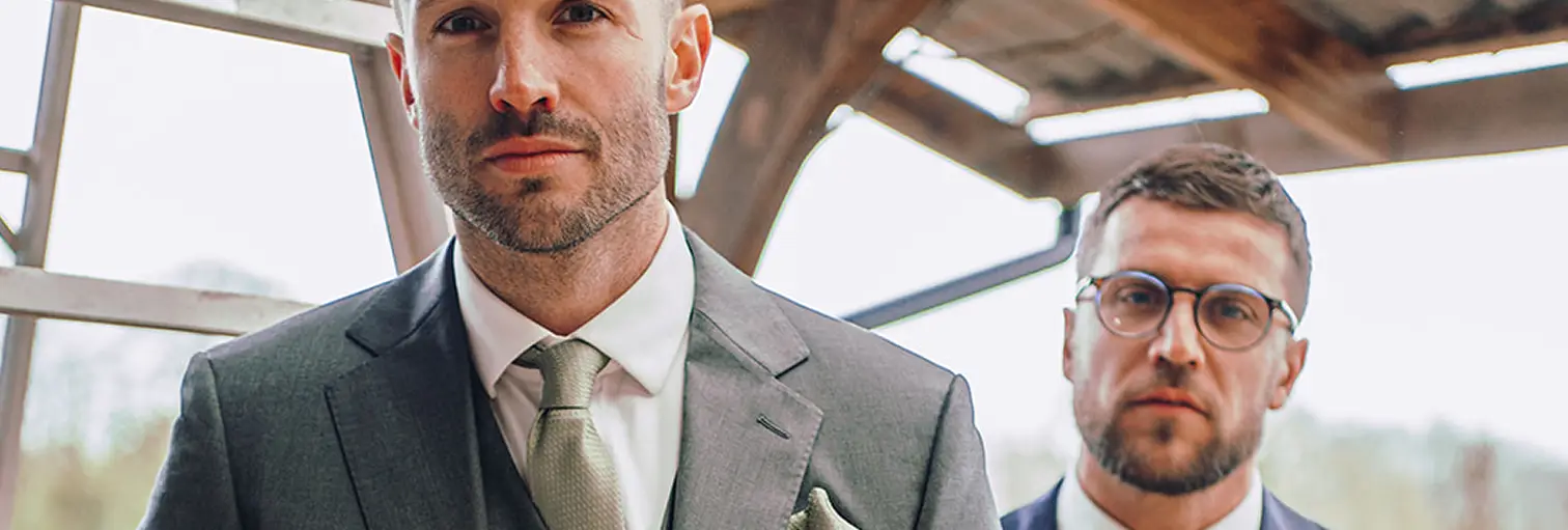 To menn i dress ser rett mot kamera i et halvåpent bygg med tretak og metallstrukturer. Den ene mannen har grå dress med slips og lommetørkle, den andre mørk dress med hvit skjorte og runde briller. Stemningen er formell og selvsikker.