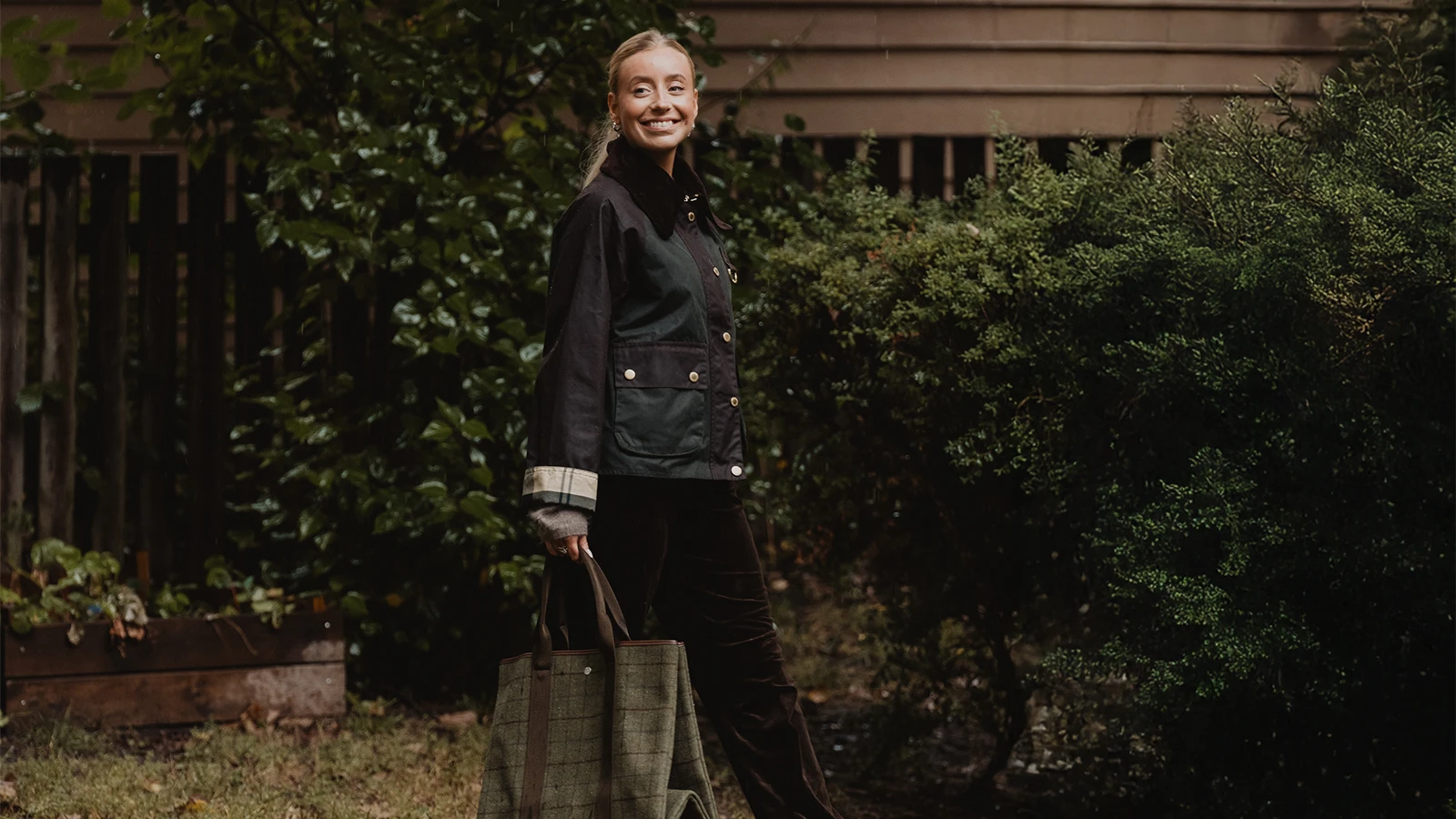 Smilende dame med bag iført mørke klær foran mørkegrønne busker