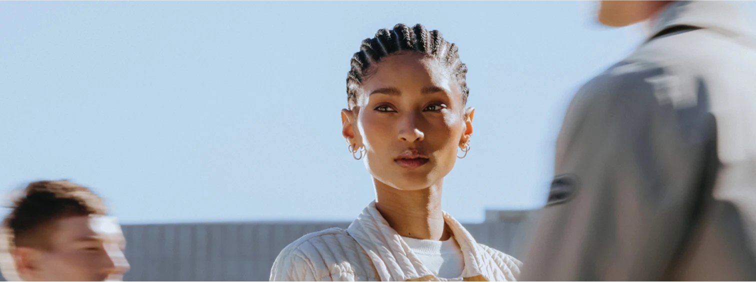 Kvinne med fletter og øreringer ser rett frem mot kamera, mens en uskarp person står i forgrunnen til høyre