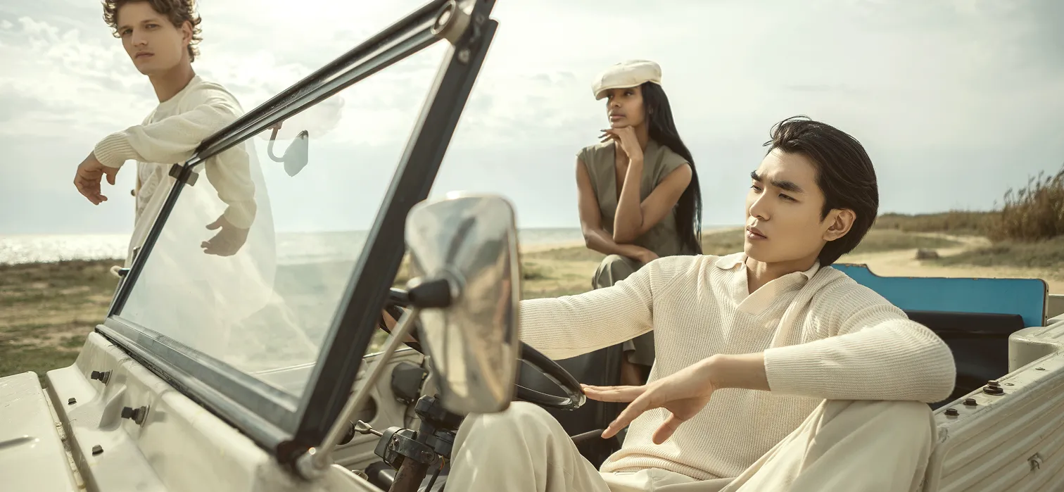 Tre stilige unge voksne ved en vintage cabriolet ved kysten – én sitter avslappet bak rattet, én står lent mot bilen, og én sitter tankefullt i bakgrunnen med hav og himmel i bakgrunnen.