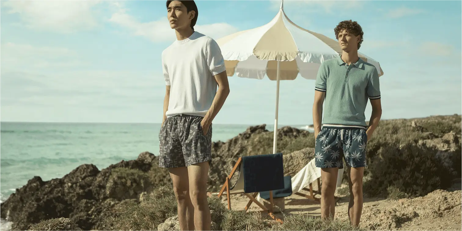 To unge menn står ved kysten iført sommerklær og badeshorts