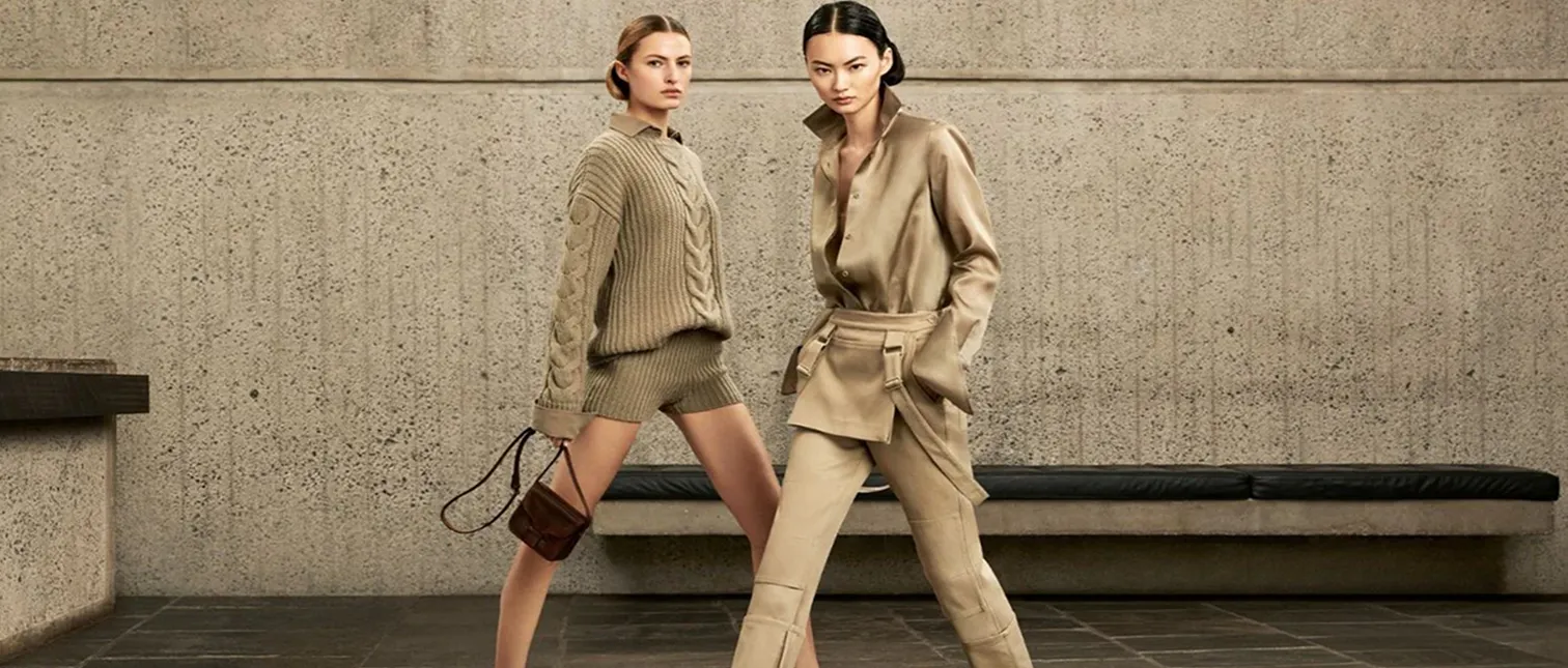 To kvinner poserer selvsikkert i sofistikerte, jordtonede antrekk foran en betongvegg. Den ene har på seg en strikket genser med shorts og holder en liten veske, mens den andre bærer et elegant satengskjorte- og bukseantrekk med brede mansjetter og beltedetaljer.