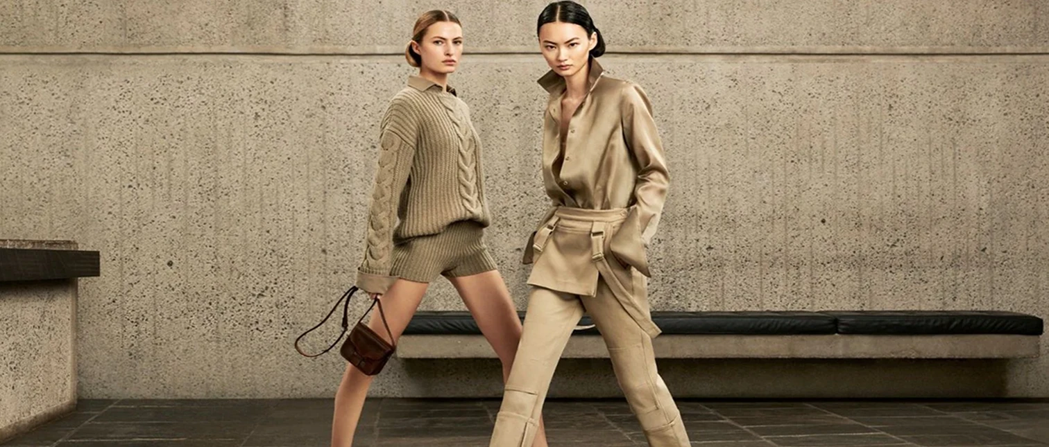 To kvinner poserer selvsikkert i sofistikerte, jordtonede antrekk foran en betongvegg. Den ene har på seg en strikket genser med shorts og holder en liten veske, mens den andre bærer et elegant satengskjorte- og bukseantrekk med brede mansjetter og beltedetaljer.