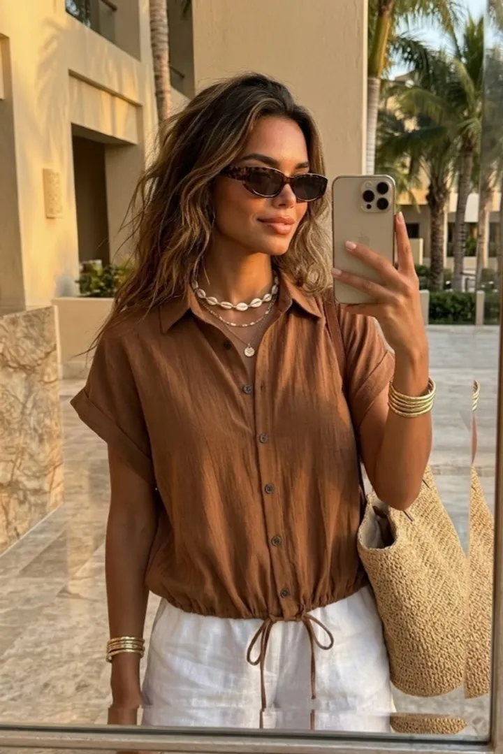 Woman wearing a brown drawstring summer blouse with white linen pants, layered gold jewelry, sunglasses, and a woven tote bag in a warm luxury resort setting.