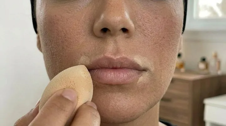 Makeup sponge blending concealer around the lips to create a clean base for a diffused wine lip makeup tutorial.