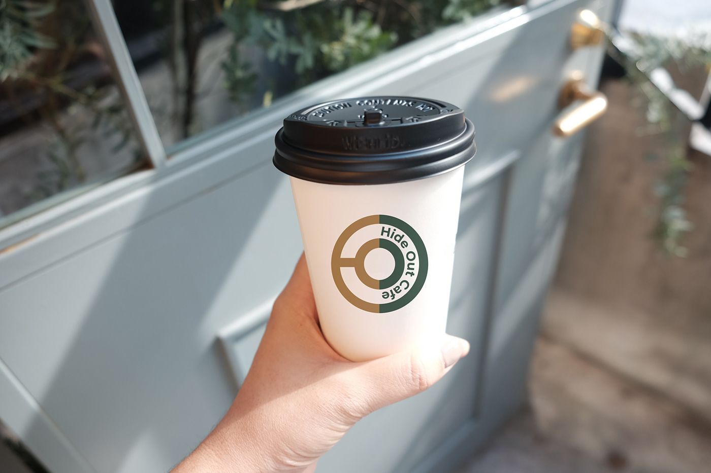 Hand holding a white takeaway cup with the Hide Out Cafe logo in green