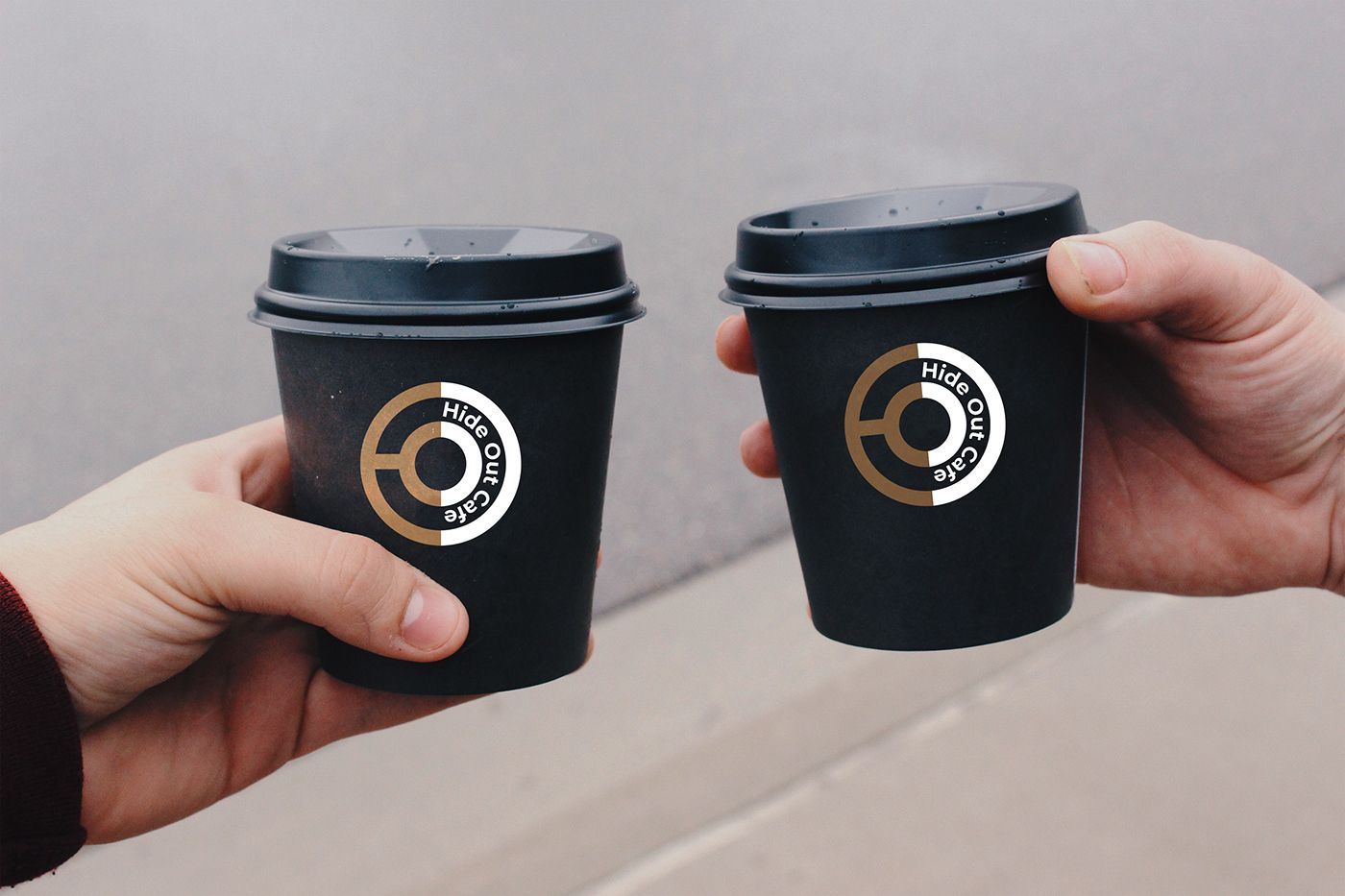 Two hands holding black takeaway cups with the Hide Out Cafe logo