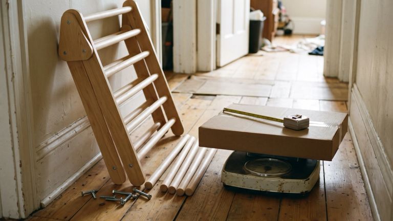 A wooden climbing triangle against a hallway wall with loose rungs on the floor, beside a flat box and tape measure on a bathroom scale