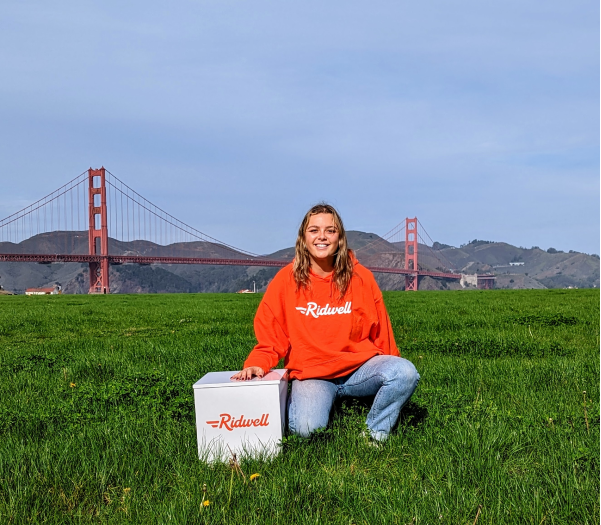 Community launcher Luna is sitting next to a Ridwell Bin wearing an Orange Ridwell sweater on Chrissy Field. In the background is the Golden Gate Bridge.