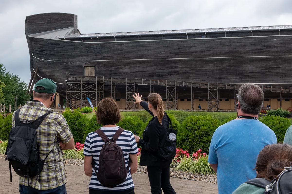 Inspire guide leads guests on a tour outside the Ark Encounter, pointing toward the life-size Noah's Ark as the group listens through wireless headsets.
