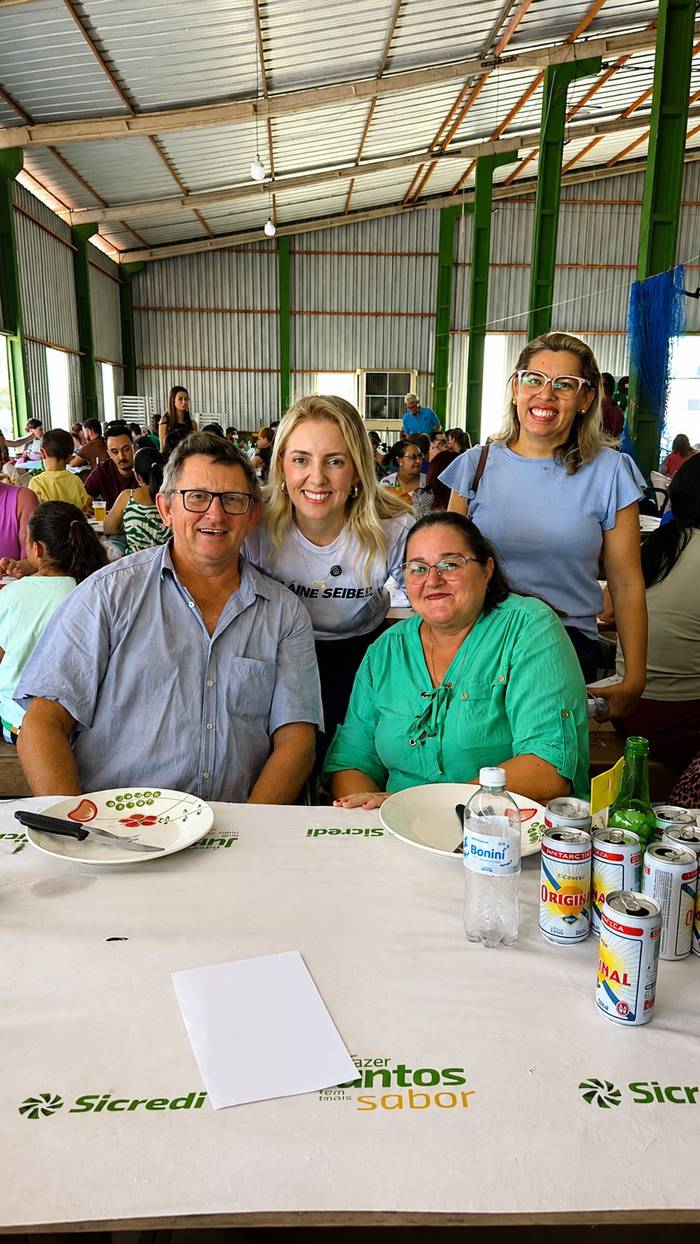 Elaine sentada à mesa entre um casal de moradores num evento comunitário.