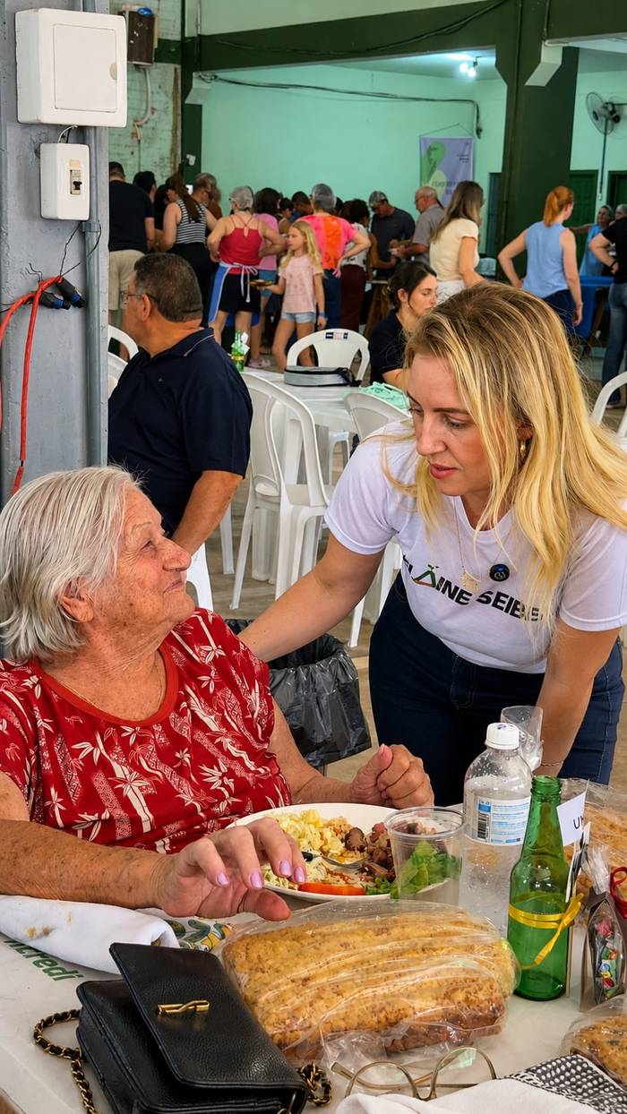 Elaine agachada, de mãos dadas com uma senhora sentada, num almoço comunitário.