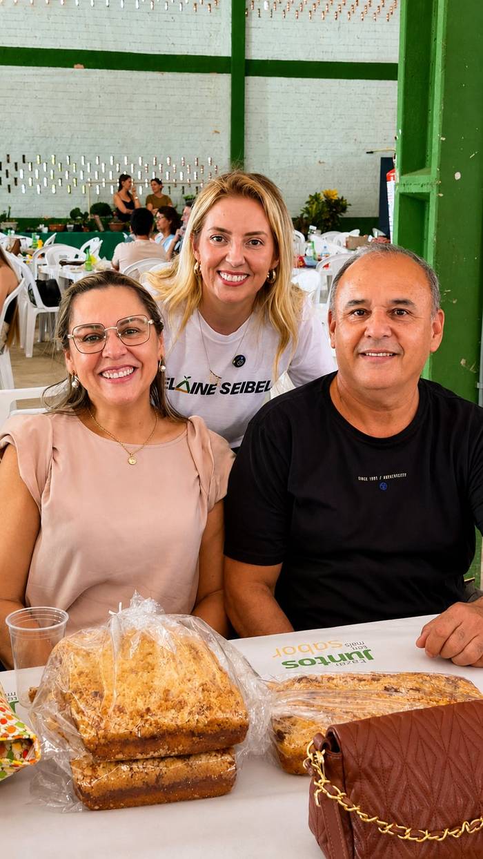 Elaine ao lado de um casal, todos sorrindo, num evento.
