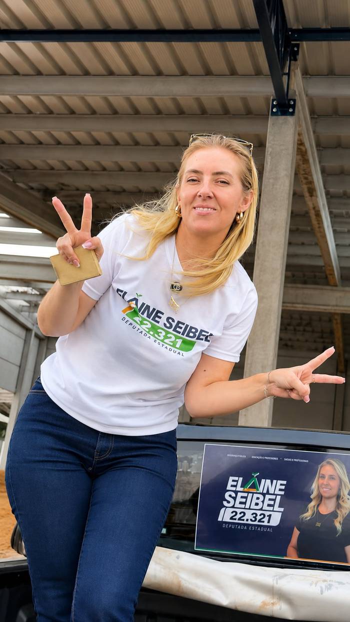Elaine com a camiseta da campanha, fazendo sinal de vitória, num galpão de armazenagem de grãos.