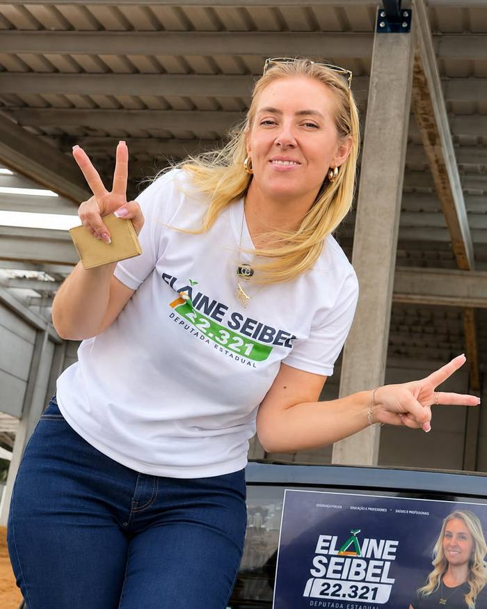 Elaine com a camiseta da campanha, fazendo sinal de vitória, num galpão de armazenagem de grãos.