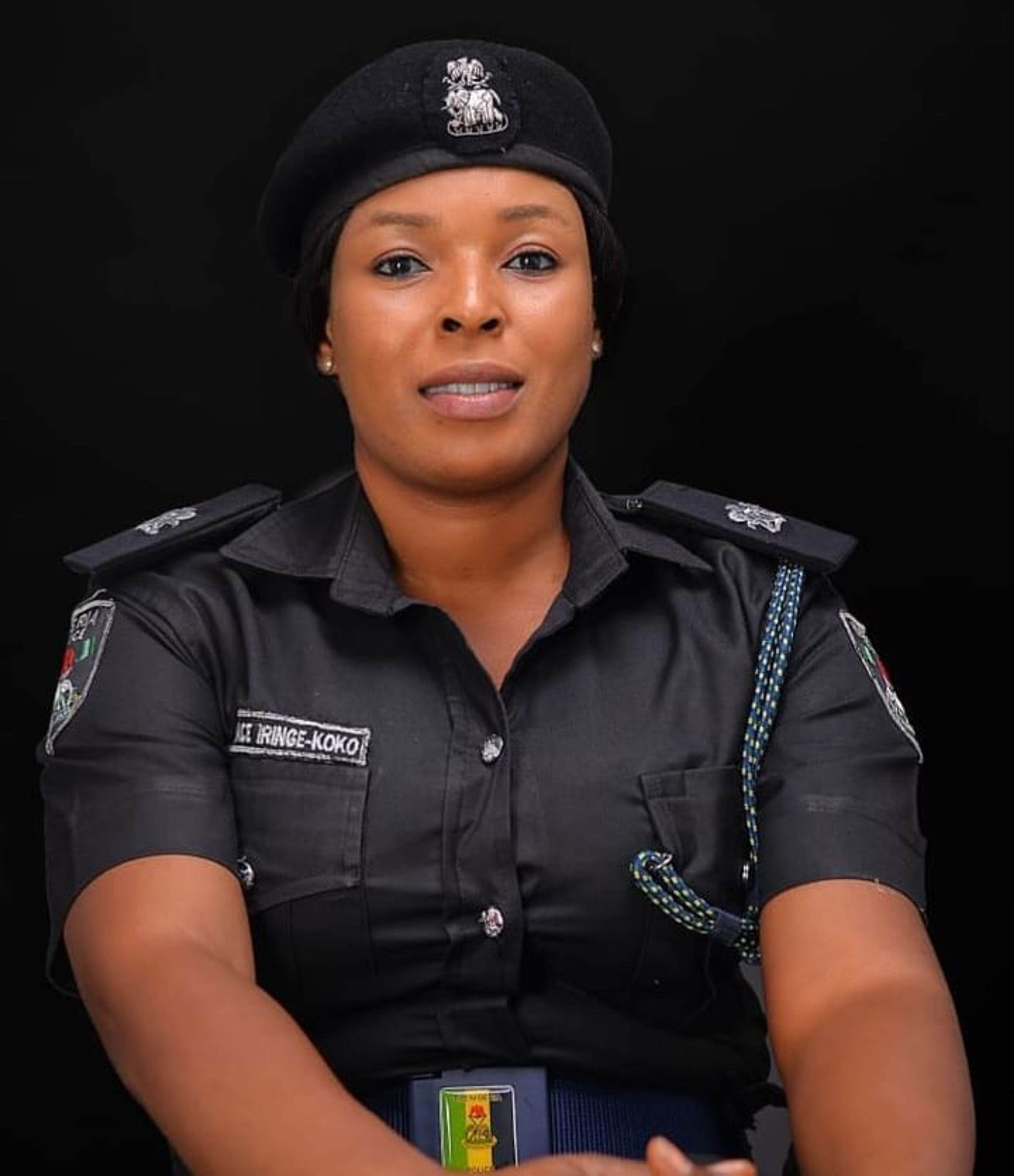 A policewoman in dark uniform and beret poses against a black background, arms folded