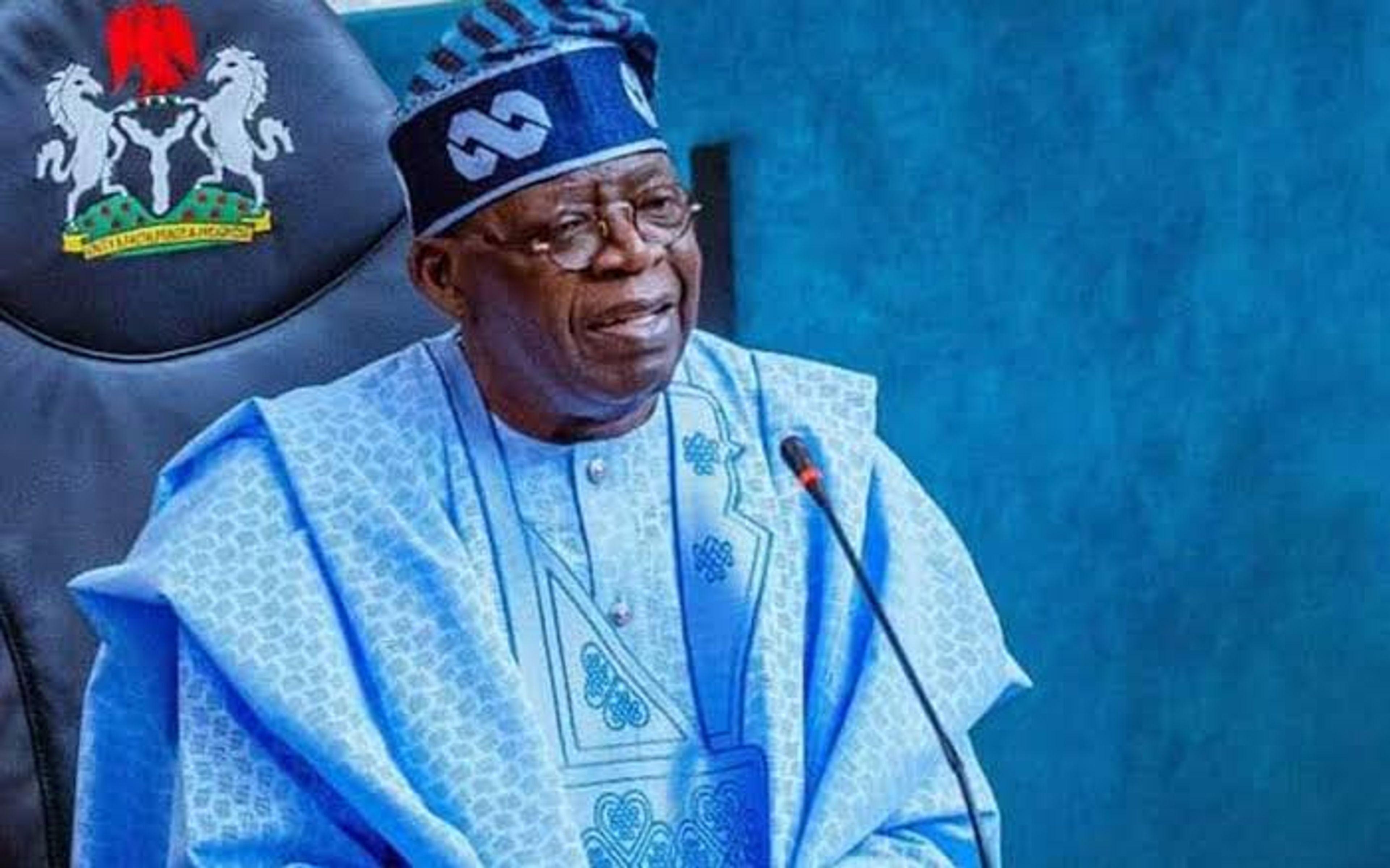 An older man in traditional blue robes and cap speaking at a microphone beside a national crest