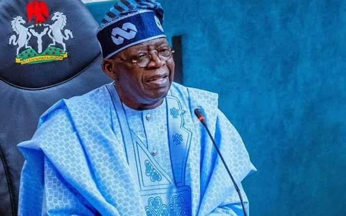 An older man in traditional blue robes and cap speaking at a microphone beside a national crest