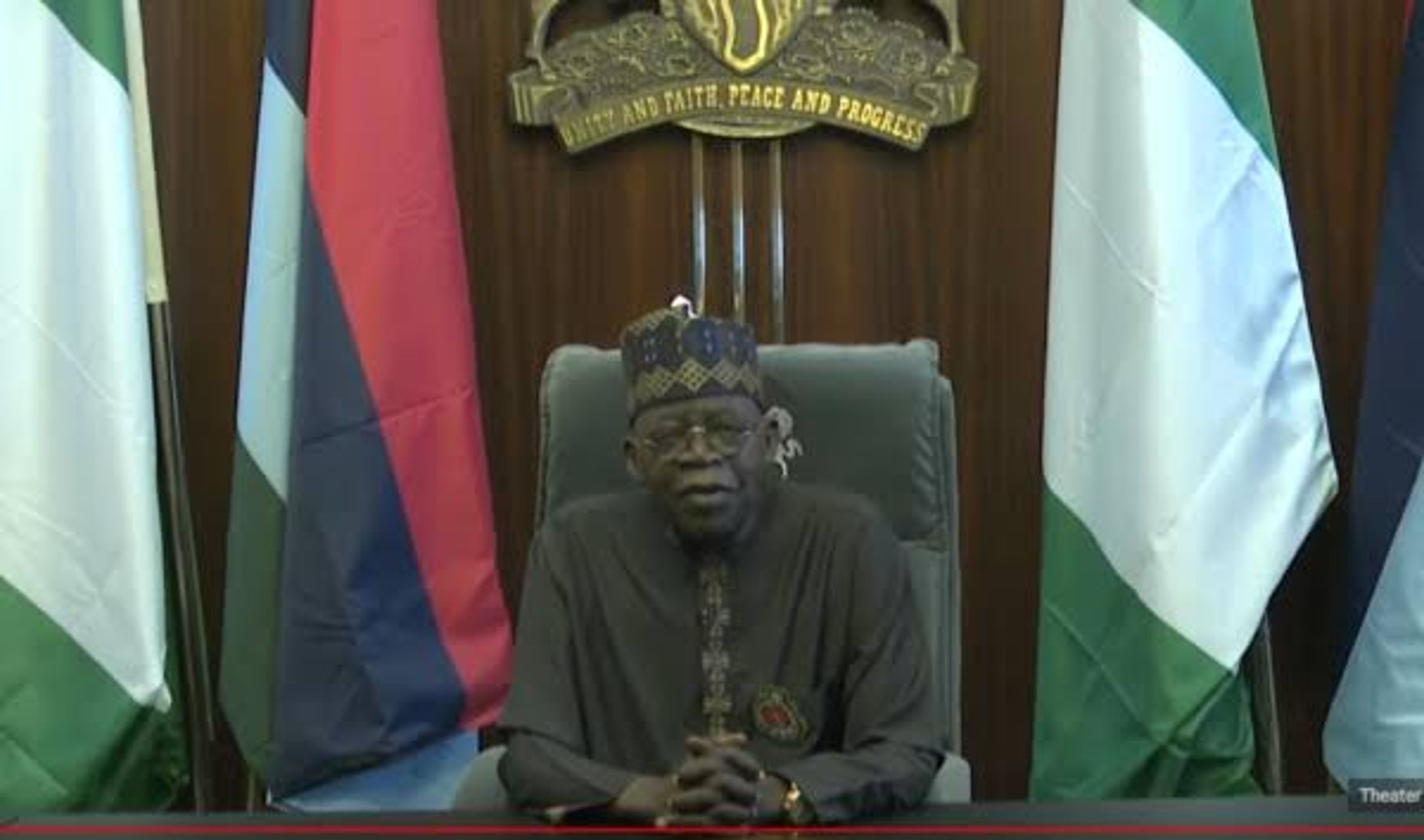 A man in a cap and robe seated before national flags and a national coat of arms