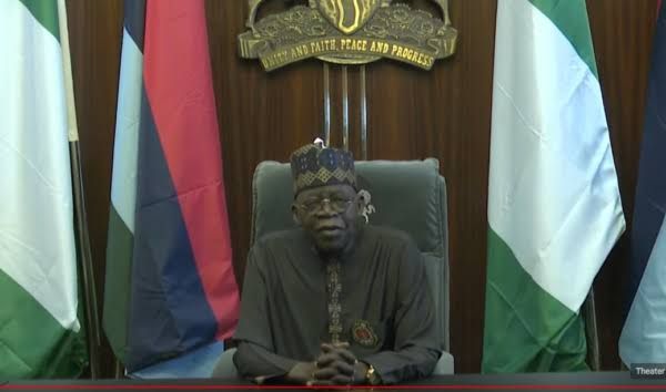 A man in a cap and robe seated before national flags and a national coat of arms