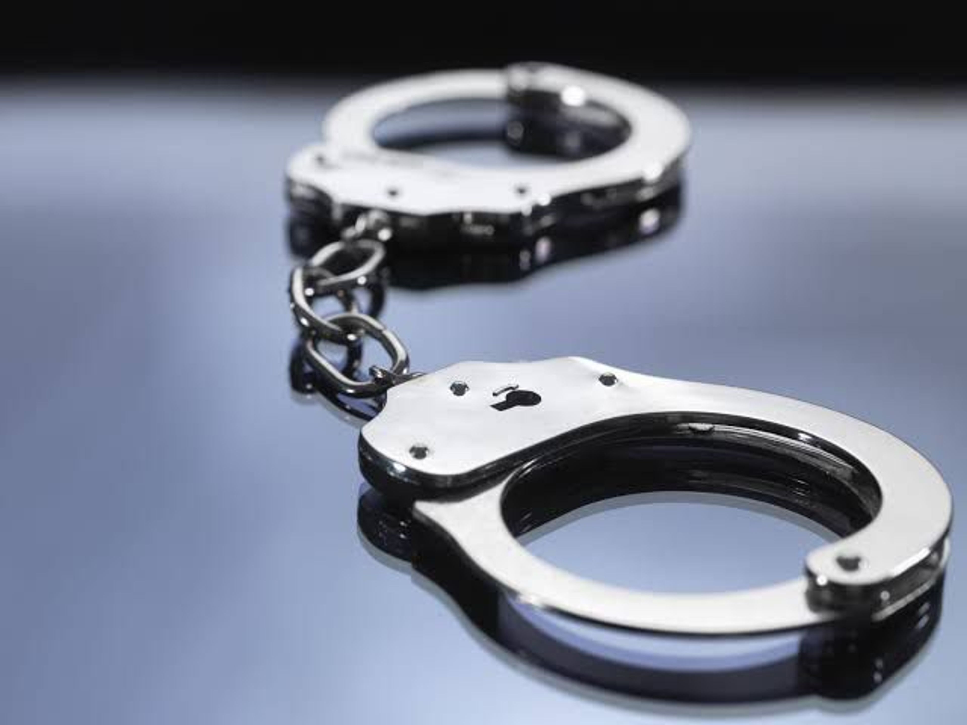 A pair of open metal handcuffs linked by a chain, resting on a reflective grey surface
