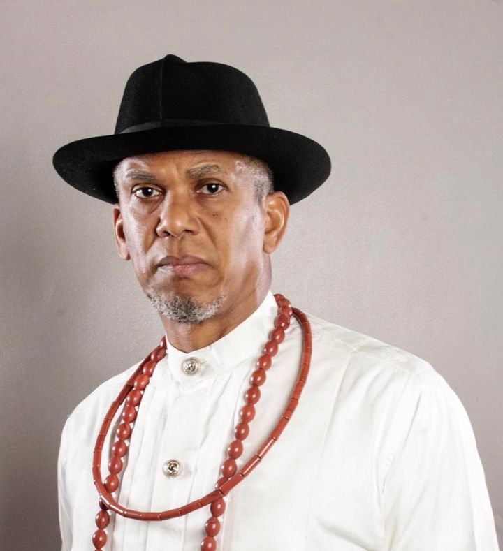 A man in a black fedora and white shirt with red coral bead necklaces against grey backdrop