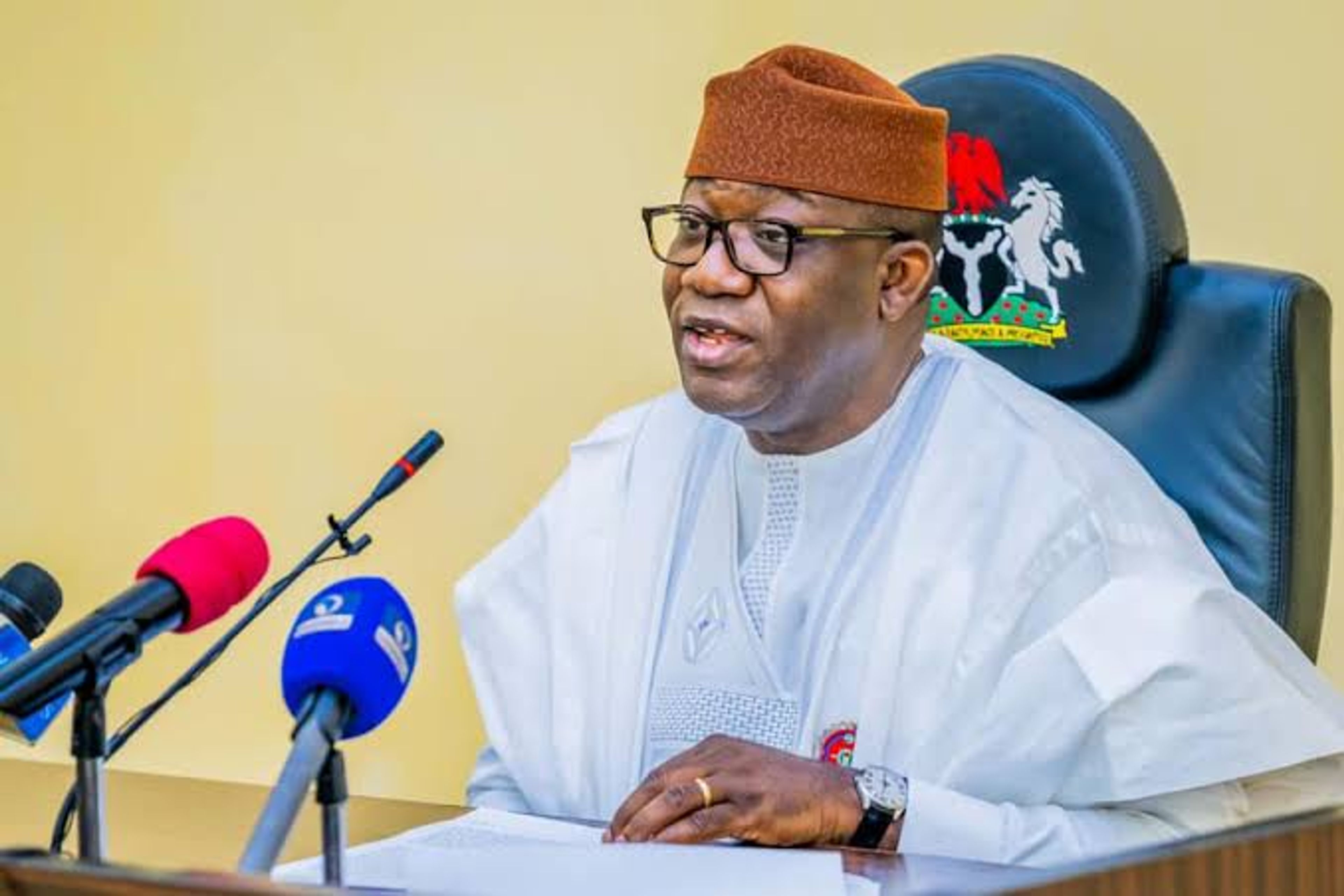 Former Ekiti State Governor, Dr. kayode Fayemi, wearing a white traditional agbada and a brown cap, speaking at a desk into multiple microphones during a press event.