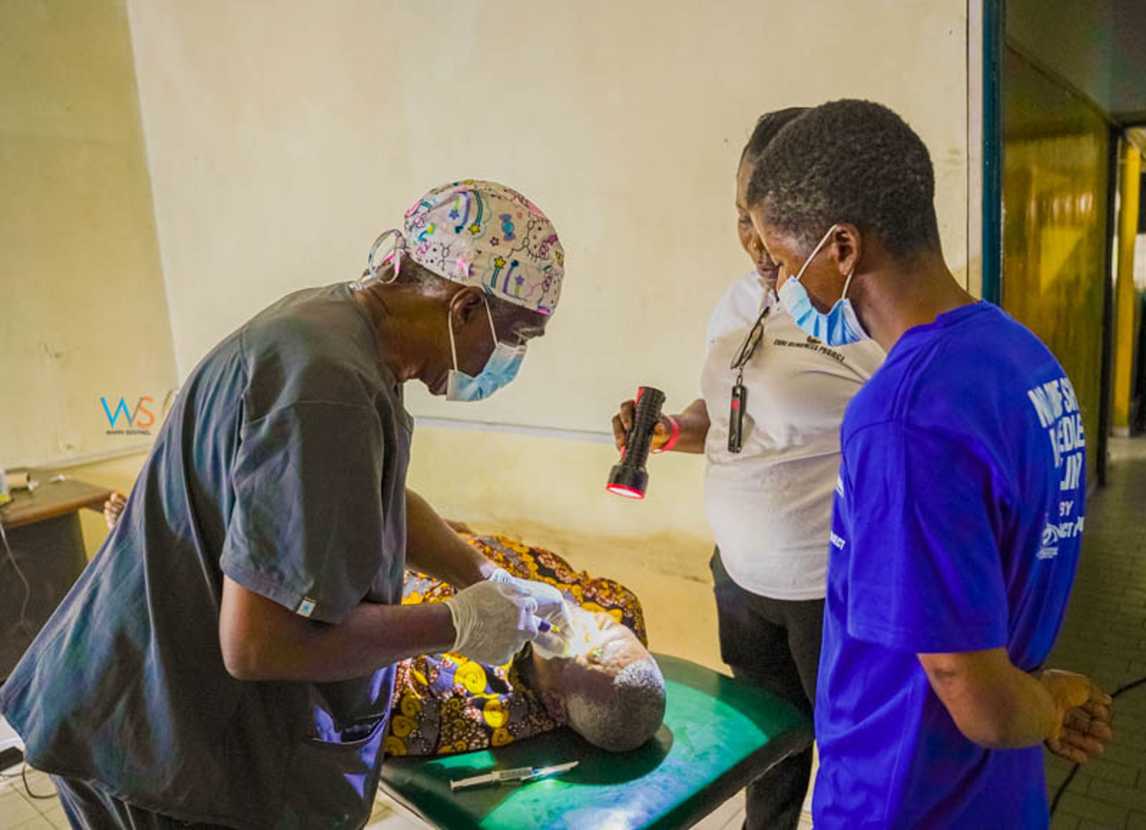 A masked surgeon treats a patient's eye while an assistant holds a torch in a clinic room
