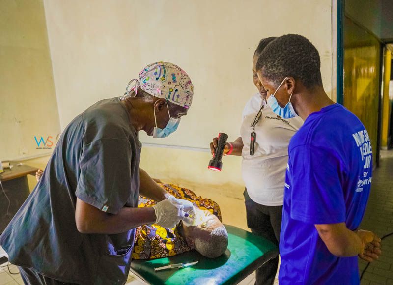 A masked surgeon treats a patient's eye while an assistant holds a torch in a clinic room