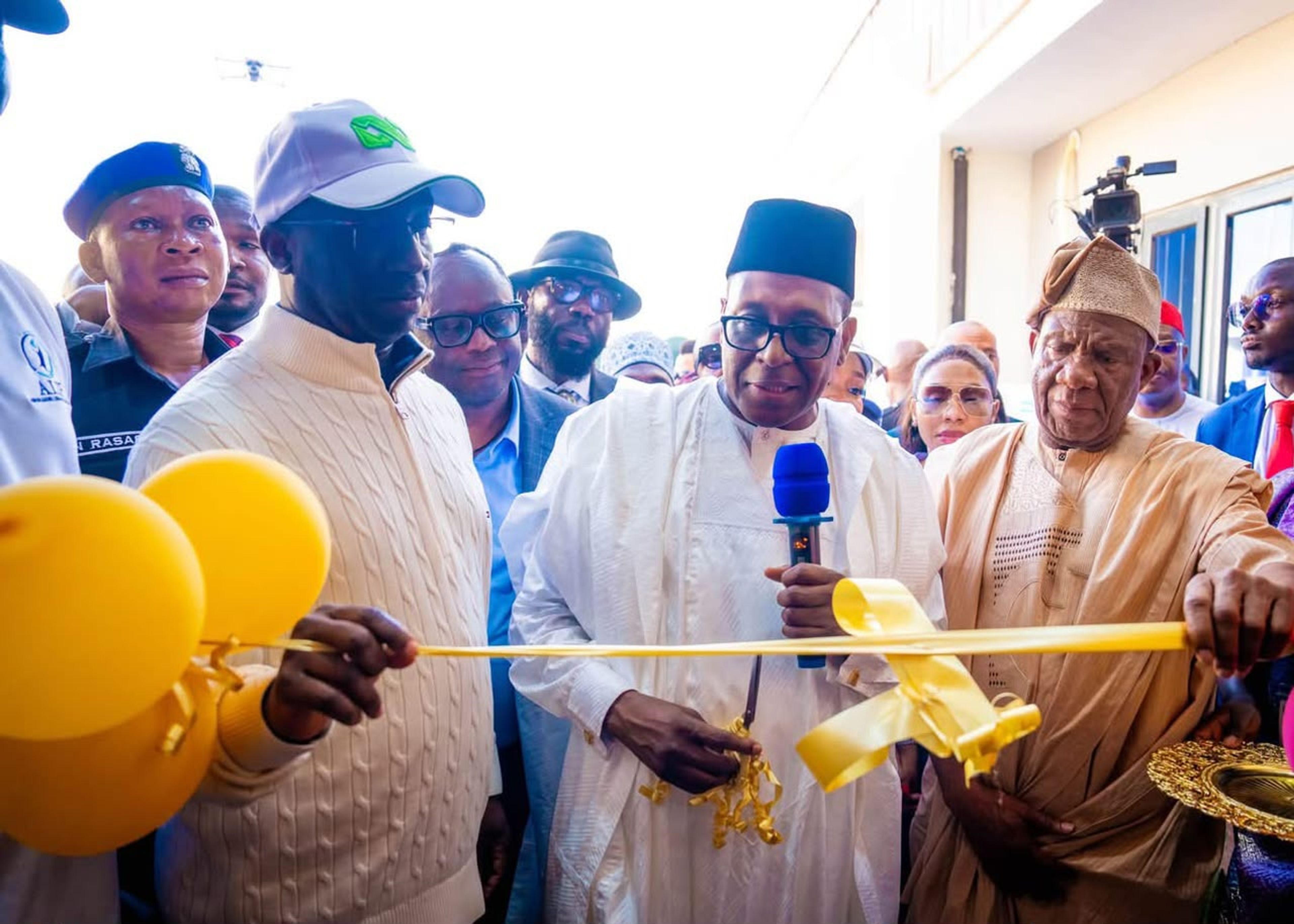A ribbon-cutting ceremony featuring several men in formal and traditional attire, including one holding a microphone while cutting a yellow ribbon, with yellow balloons visible in the foreground.