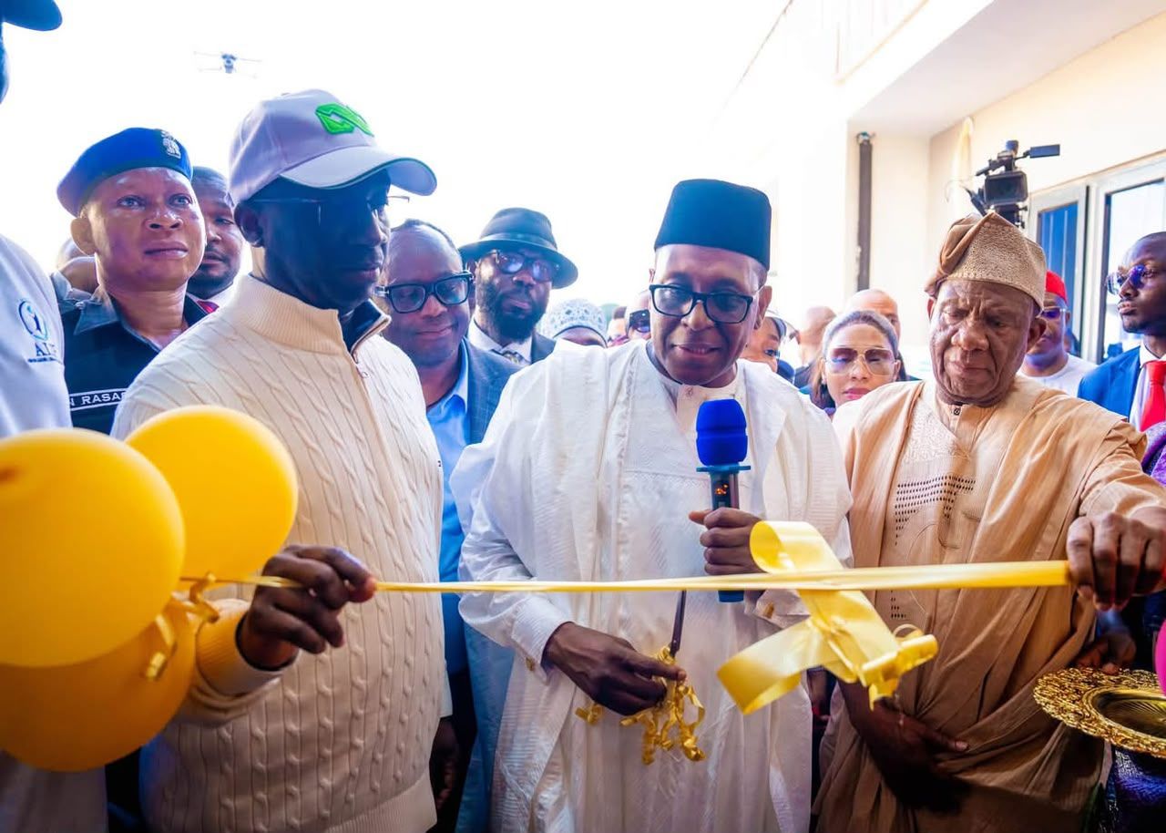 A ribbon-cutting ceremony featuring several men in formal and traditional attire, including one holding a microphone while cutting a yellow ribbon, with yellow balloons visible in the foreground.