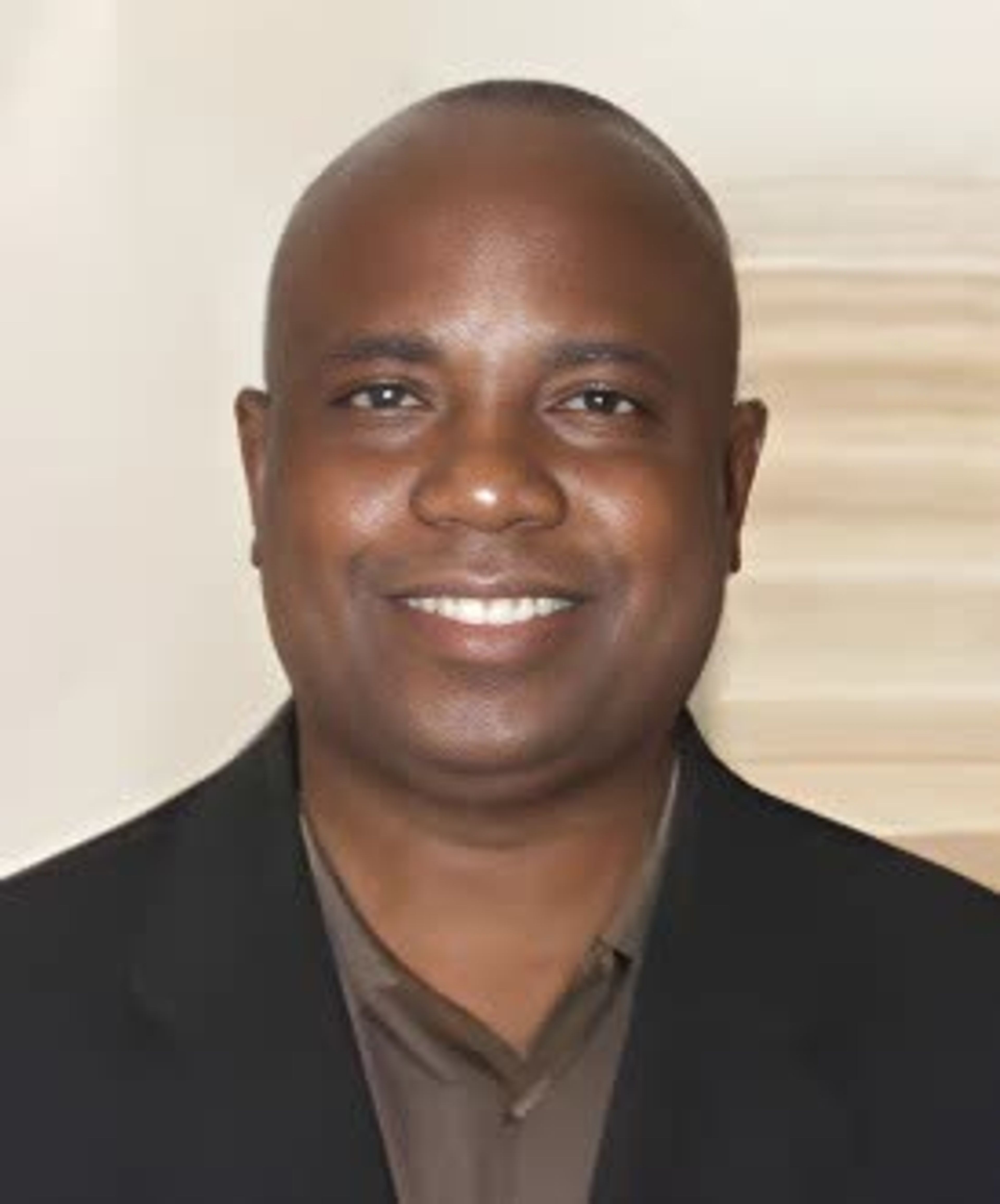 Smiling man in a dark suit jacket and brown shirt posing for a portrait against a pale background