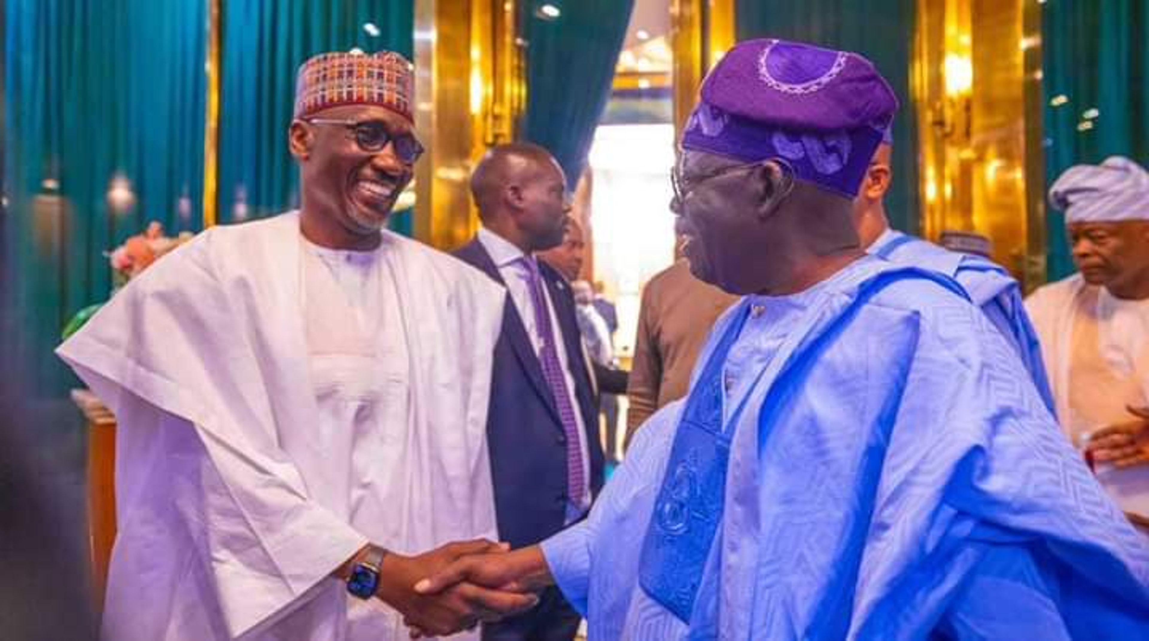 Two men in traditional attire shake hands and smile warmly at an indoor gathering with others nearby