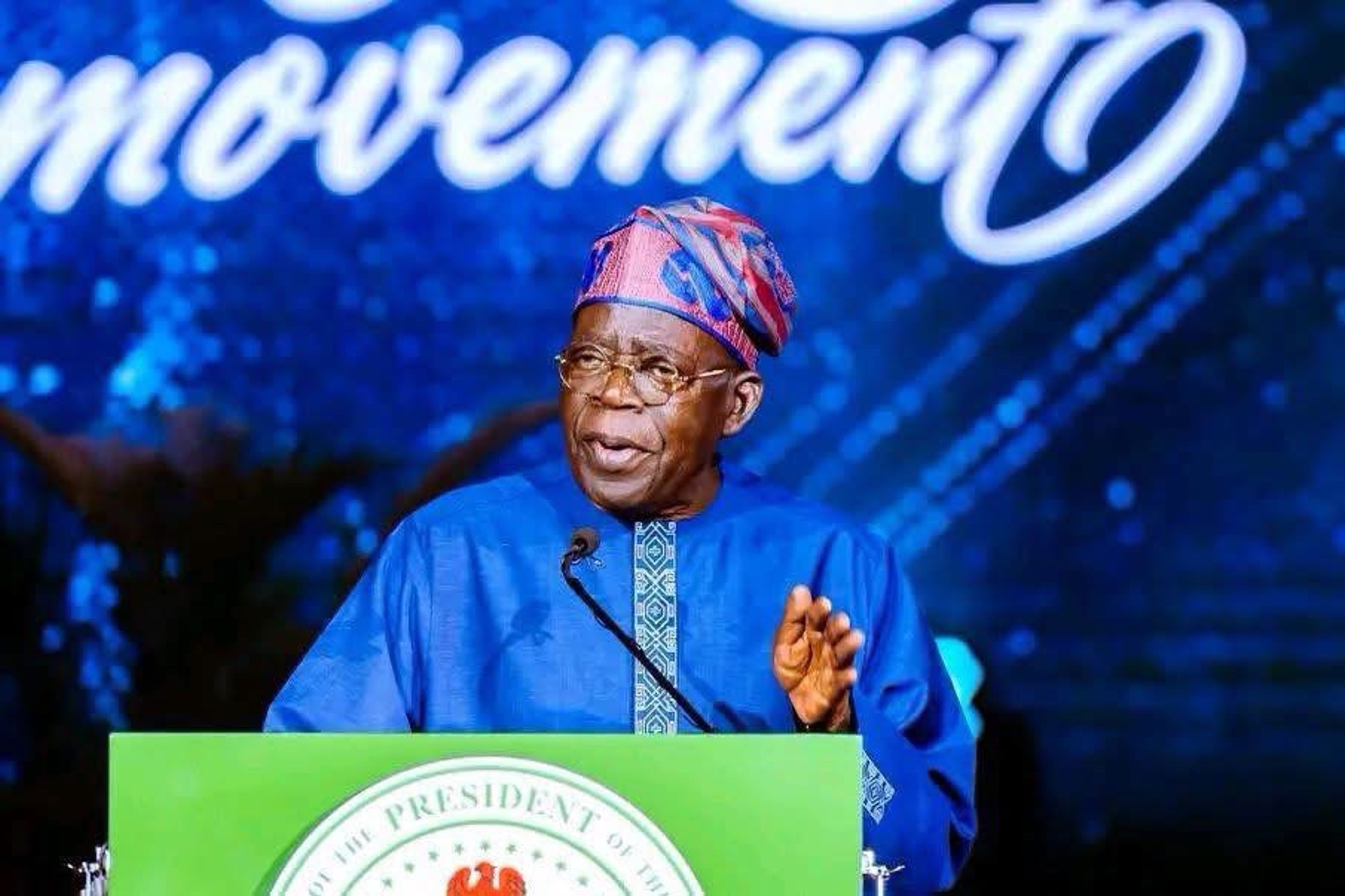 Close-up photograph of Nigerian President Bola Ahmed Tinubu speaking at a green presidential podium during a City Boy Movement event, framed against a dark blue background with white stylized text reading "movement".