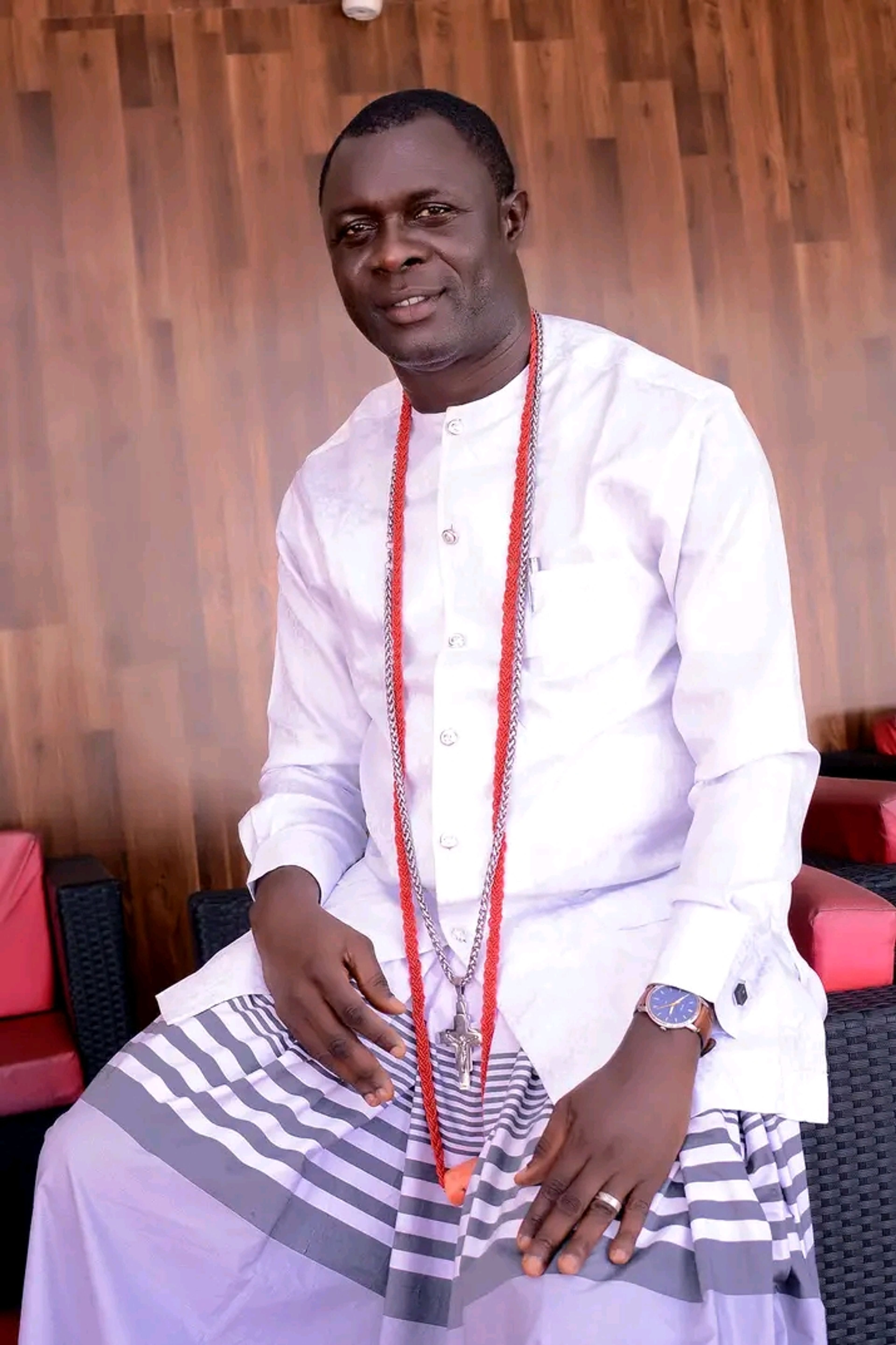 A man in white traditional attire with red beads and a cross pendant, seated indoors against wood panelling