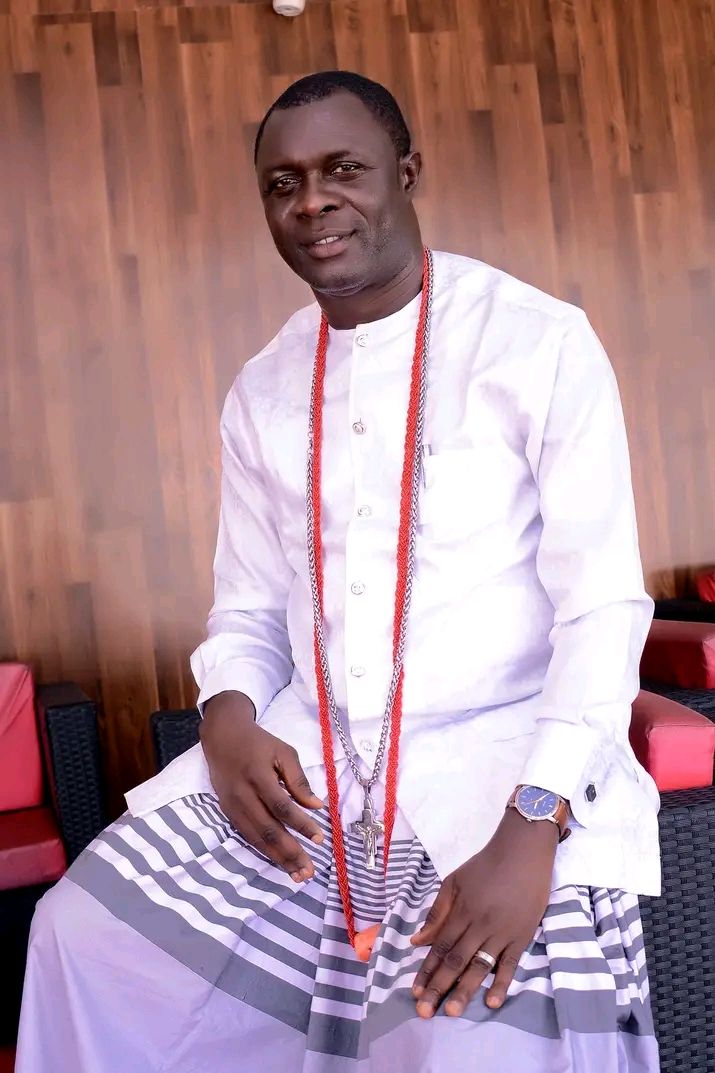 A man in white traditional attire with red beads and a cross pendant, seated indoors against wood panelling