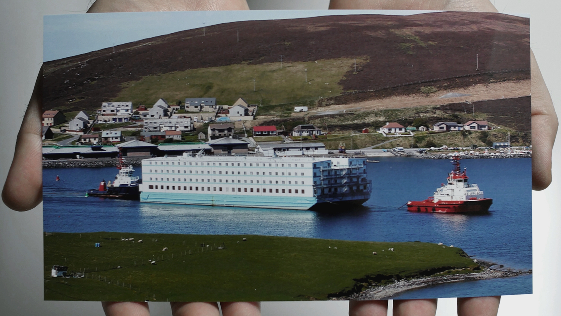 Two hands hold a photo of a barge being pulled through a strait of water.