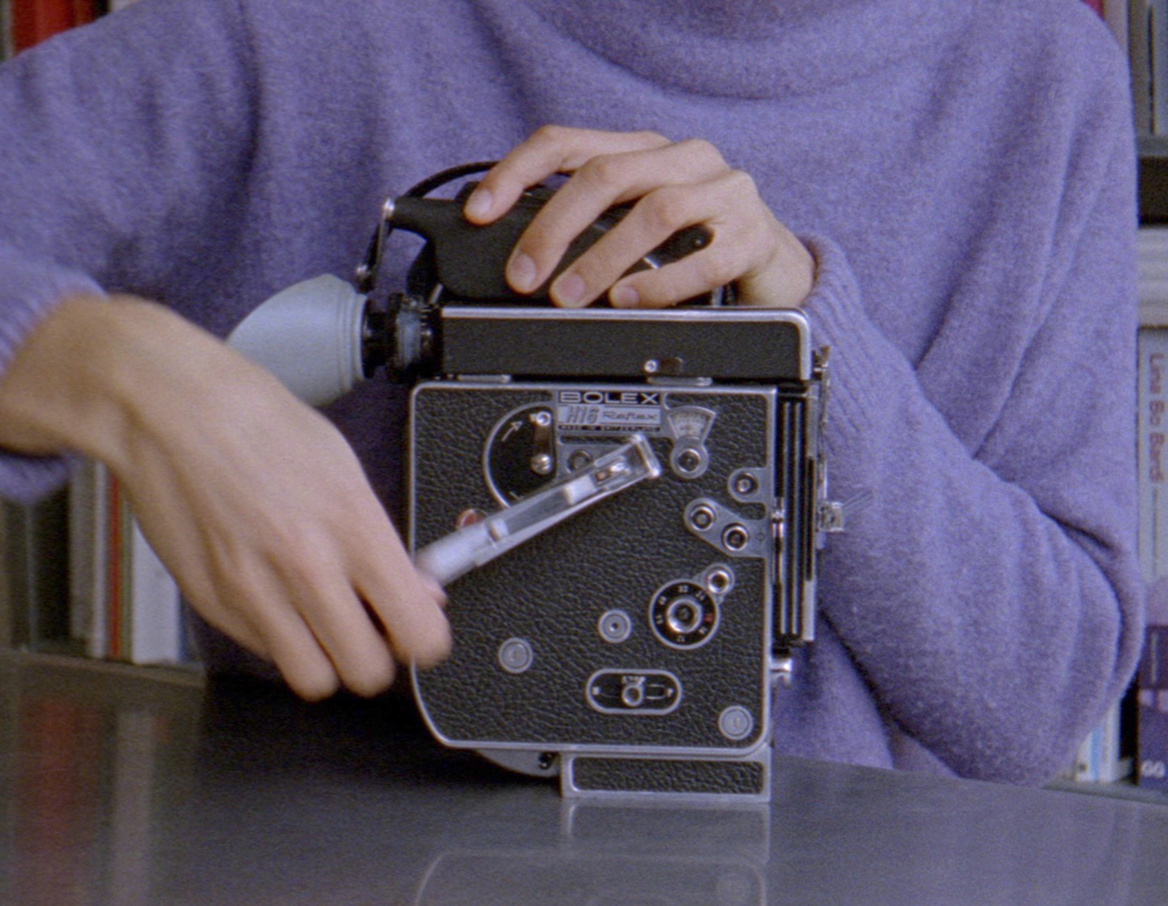 The body of a person in a lilac jumper winds up an analogue Bolex camera.