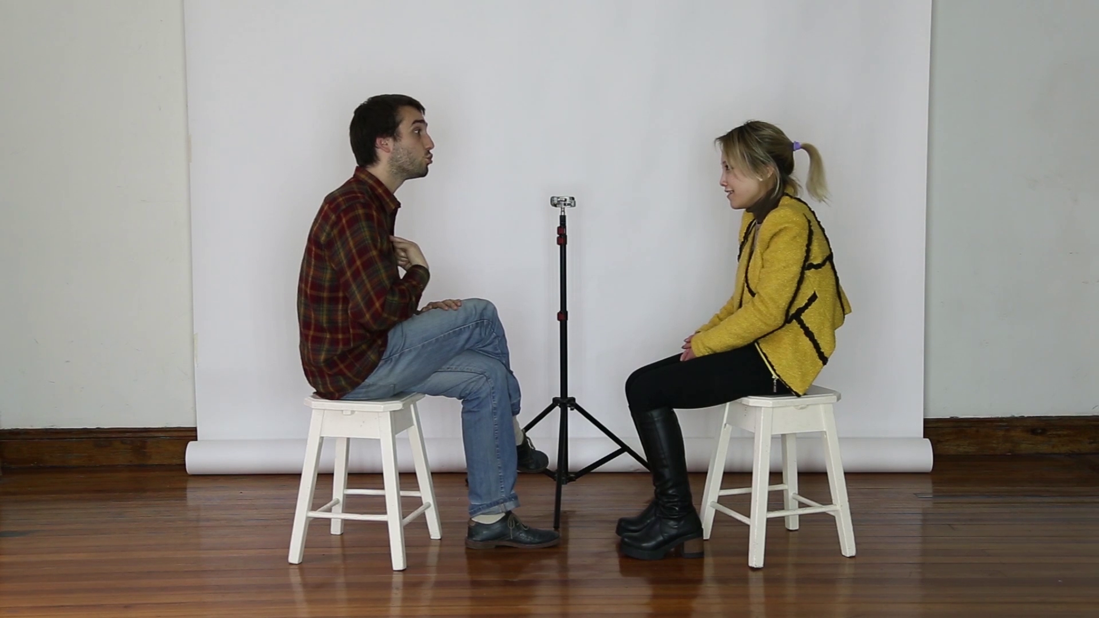 A man and woman sit on stools facing each other, with a microphone between them 