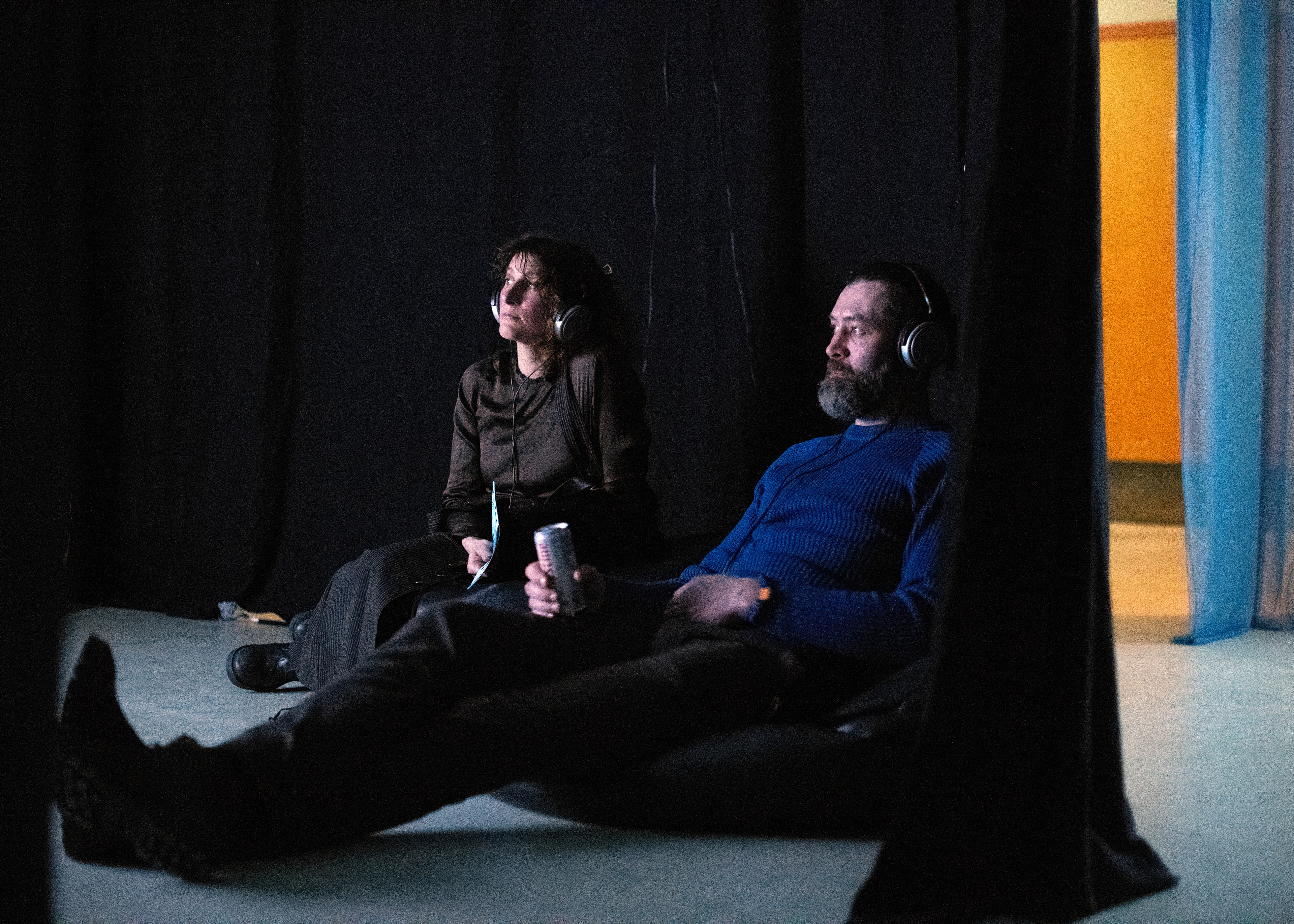 Two people sit on the floor and watch something with headphones on