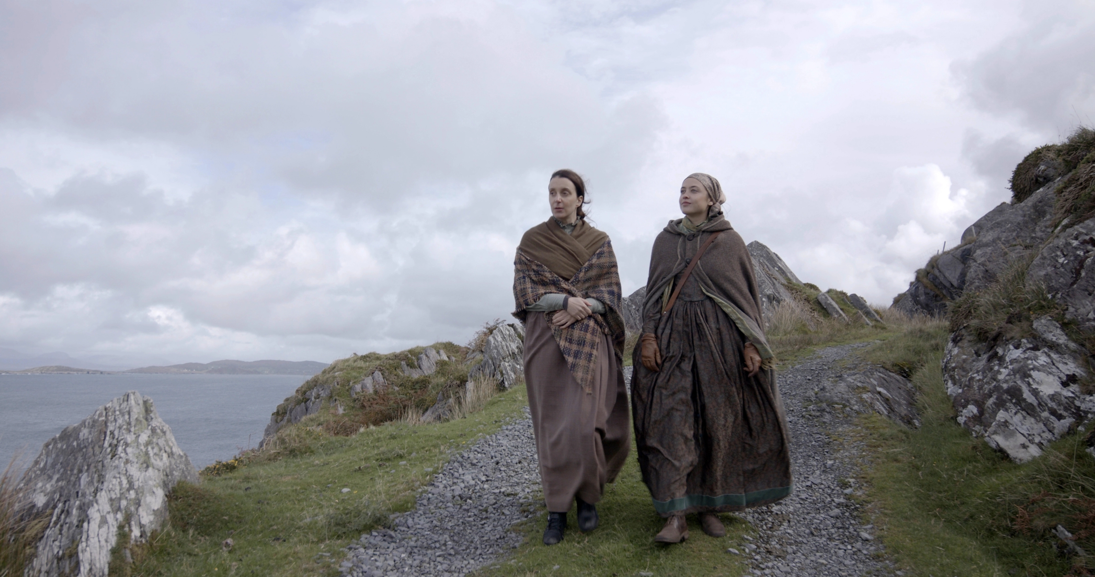 Two women in brown cloaks walk along a stone path by the sea.