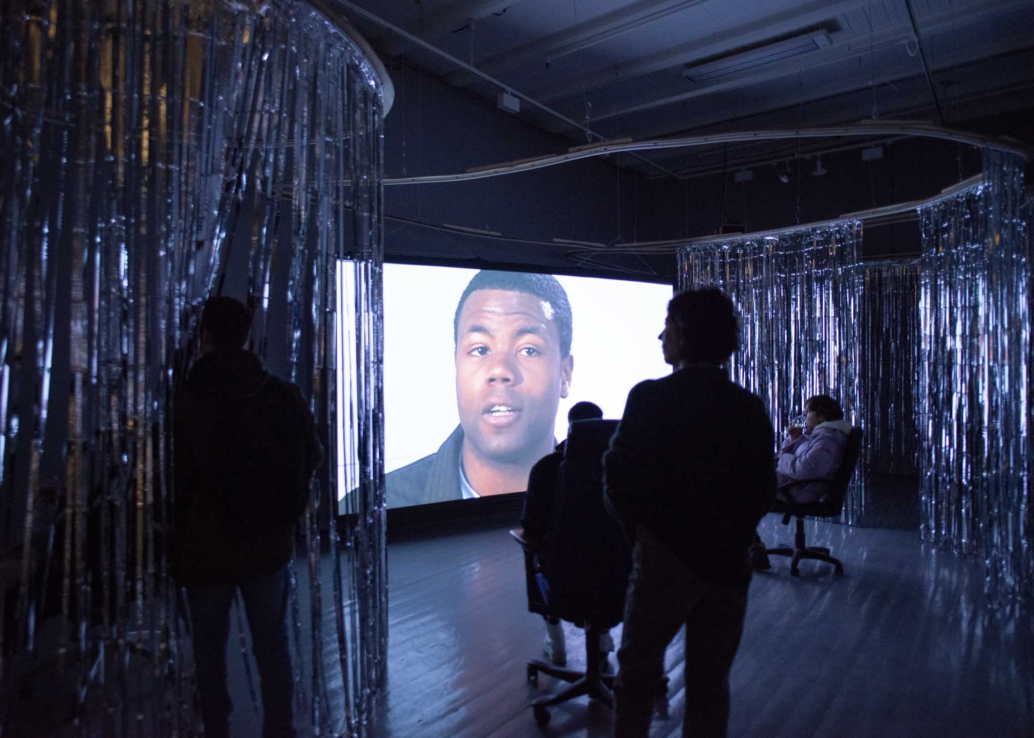 Folk sitter i kontorstoler og ser på en film i et mørkt galleri