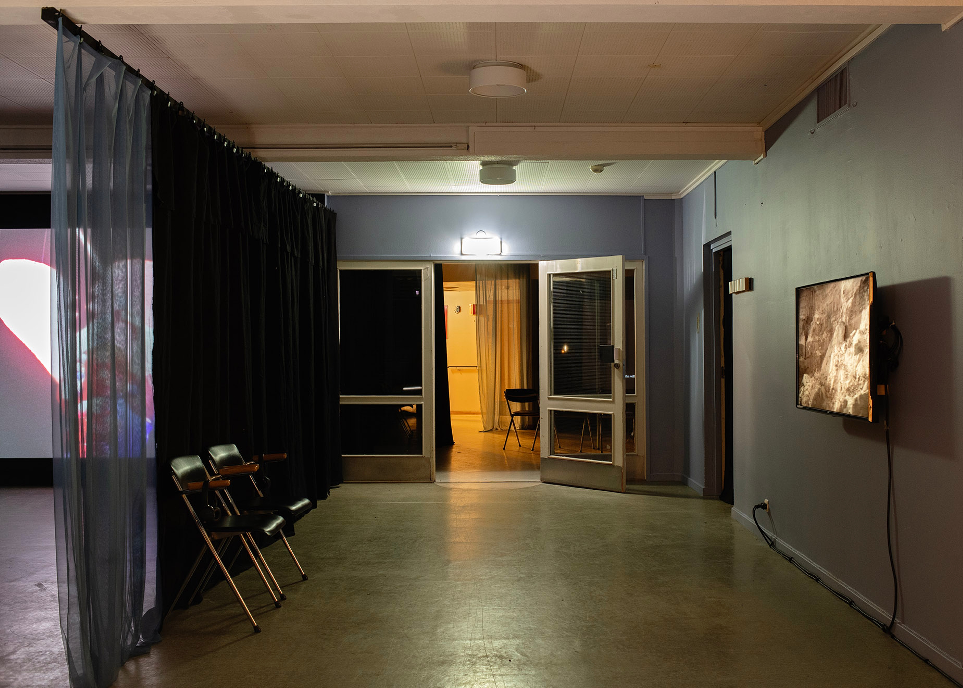 A screen on the right of a dark blue gallery. Doors at the end reveal a yellow light and a projection screen on the left is partially hidden by a curtain. 