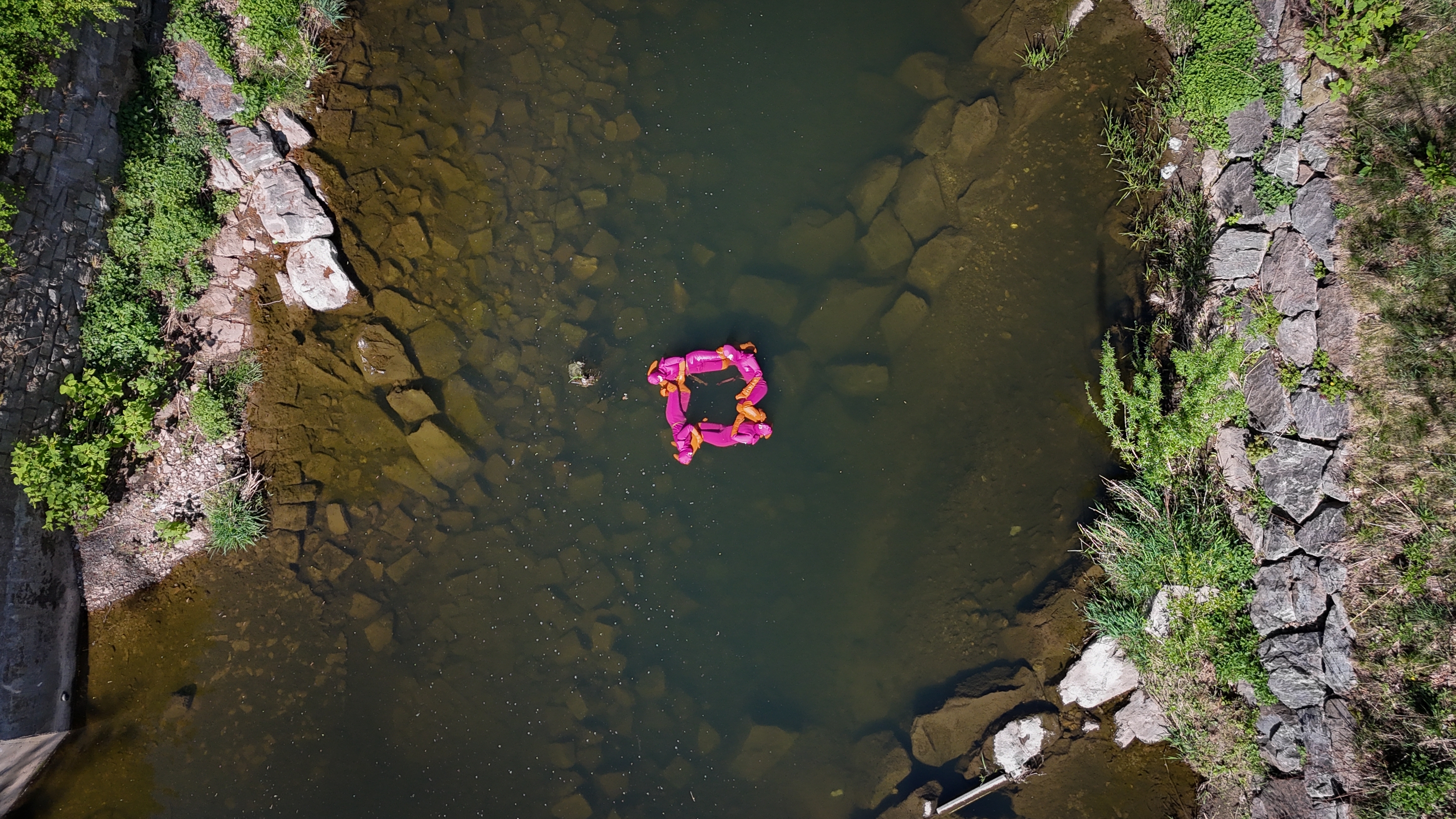 A birds eye view over a shallow river. Green foliage borders the river, and floating in the centre is a square created by 4 people floating in pink.