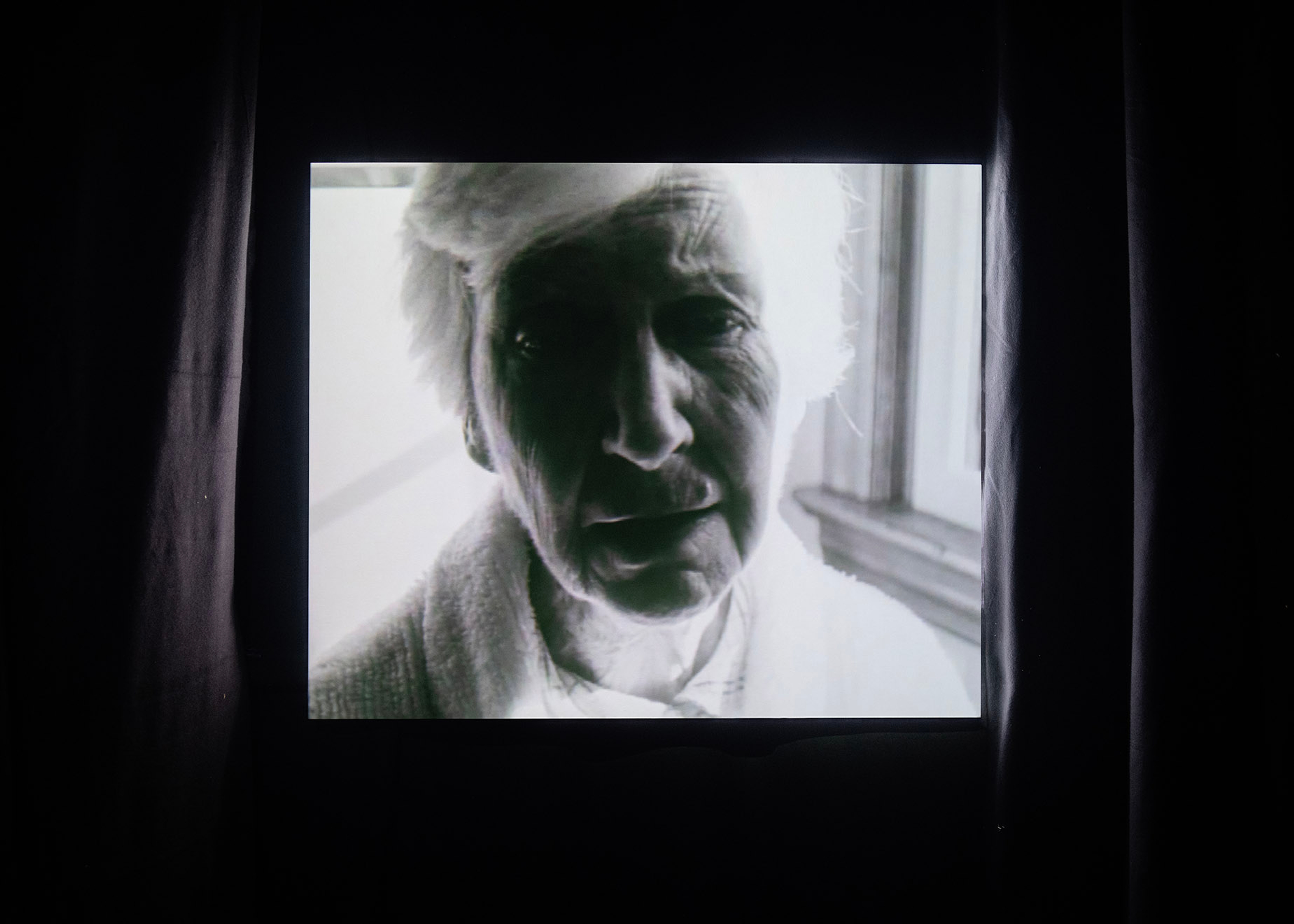 The face of an old women is projected onto a 4:3 screen in a black curtained room