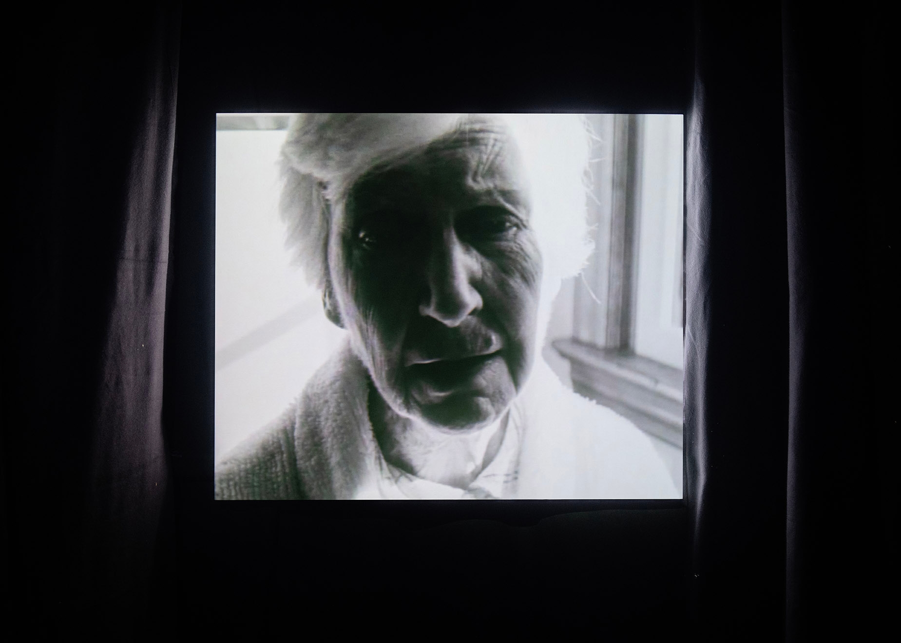 The face of an old women is projected onto a 4:3 screen in a black curtained room