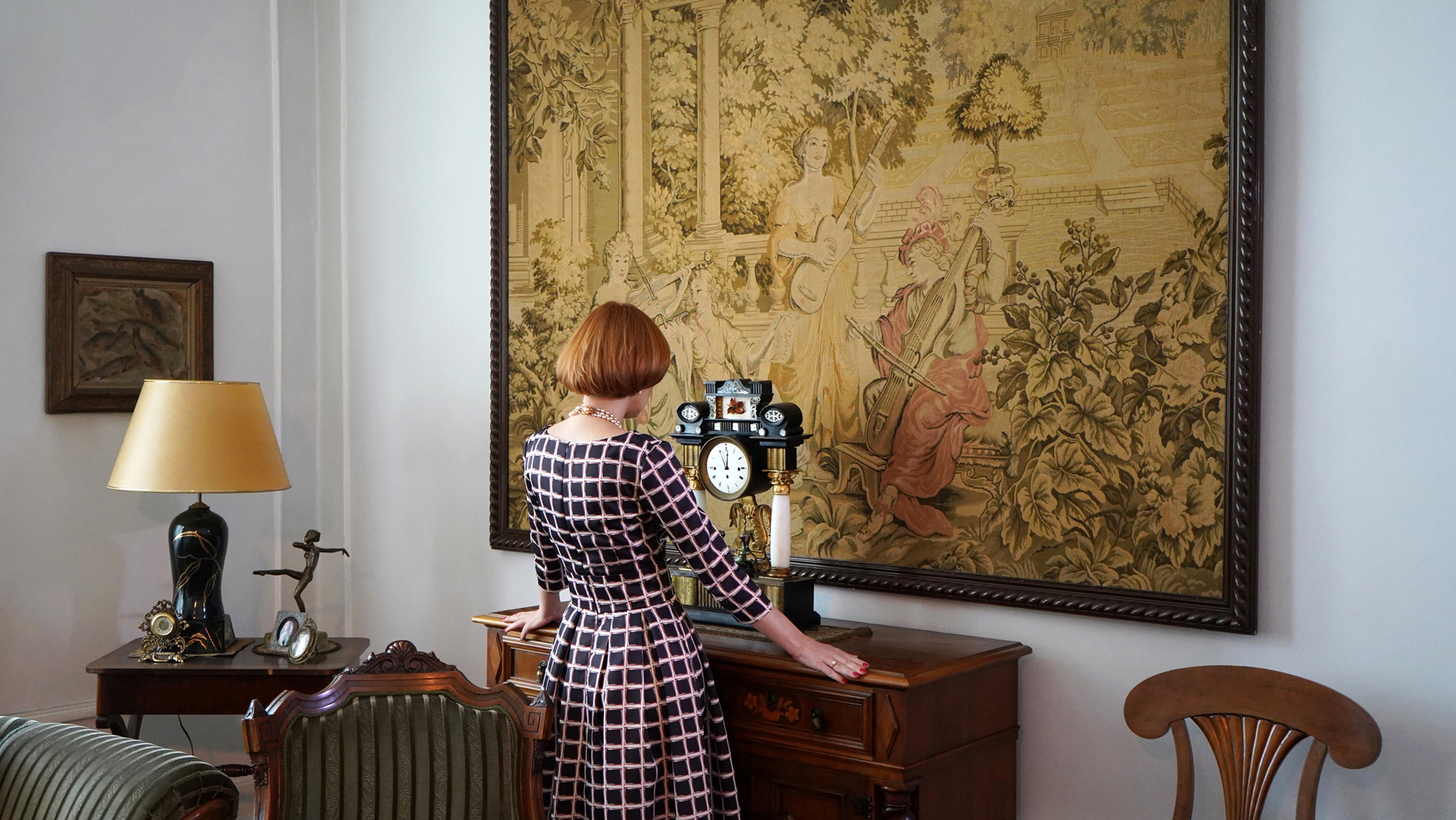 The back of a person in a dress, as they stand facing a dresser in an ornate room.